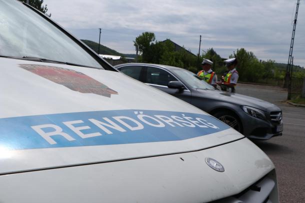 Egy rendőrautó motorháztetején olvasható a „rendőrség” felirat, háttérben az állomány két tagja egy Mercedes típusú gépjármű sofőrjének adatait kéri le.