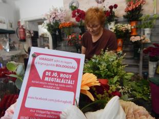 A József Attila úti virágbolt tulajdonosa készít egy csokrot, előtte a virággal egy mosolyért kampányt népszerűsítő kártya látszik.