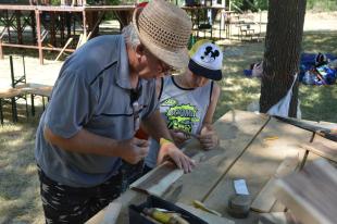 A foglalkozás vezető a fafaragás mozdulatait tanítja az egyik táborozónak.