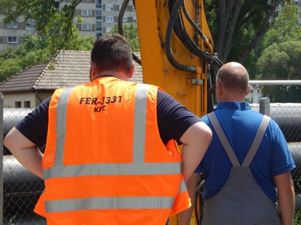 A kivitelező cég munkatársai háttal állnak a markoló gép előtt.