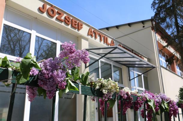 Az Ózdi József Attila Gimnázium és Kollégium főbejárata, előtte található kerítésen virágok díszelegnek.