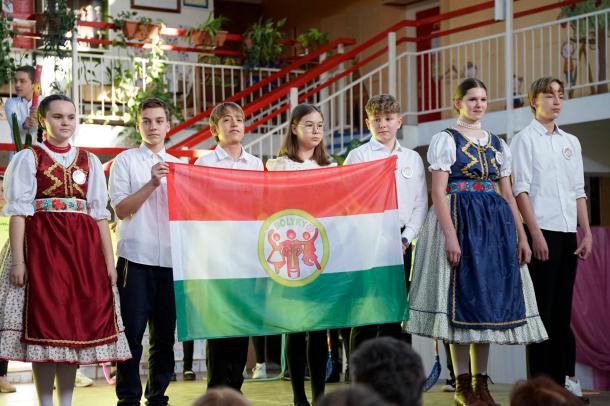 Magyar zászlót tartanak a néptáncos gyerekek, közepében az iskola logójával.