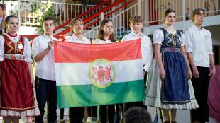 Magyar zászlót tartanak a néptáncos gyerekek, közepében az iskola logójával.